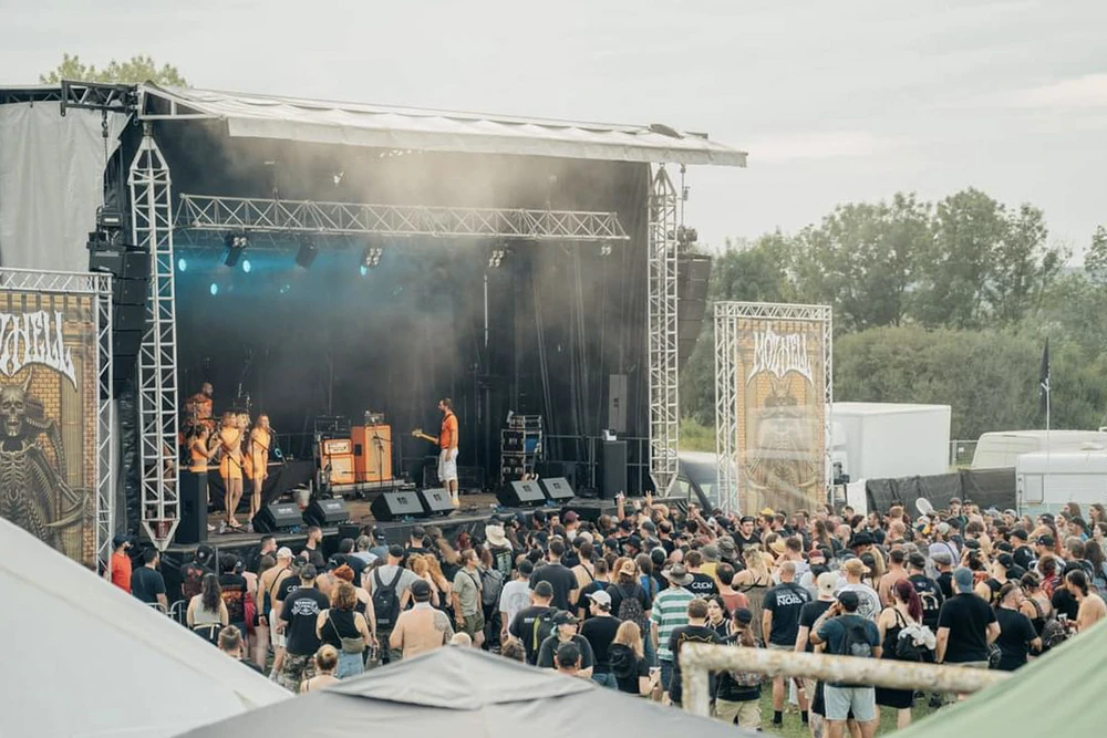 Scène extérieure de festival musical avec scène couverte, public dense debout devant performers