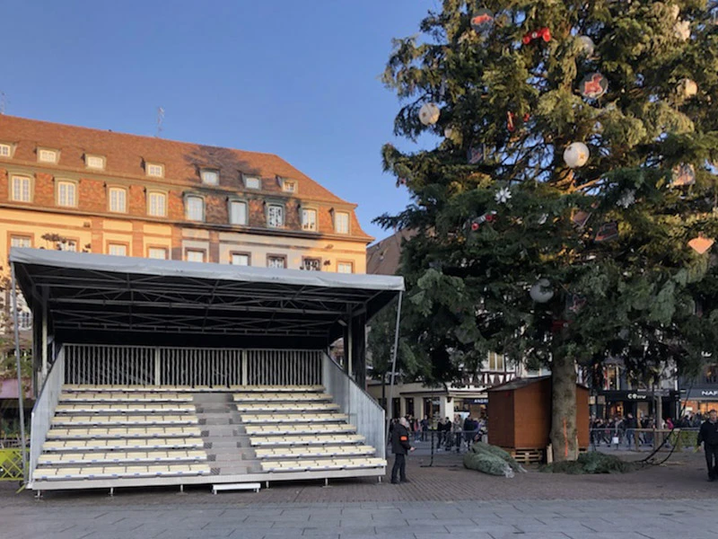 Scène extérieure d'événement musical avec scène couverte et public diversifié dans un espace urbain pavé, entourée de végétation et bâtiments résidentiels